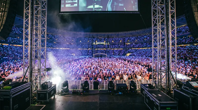 A massive stadium crowd at night, entirely illuminated by synchronized blue and purple LED wristbands.
