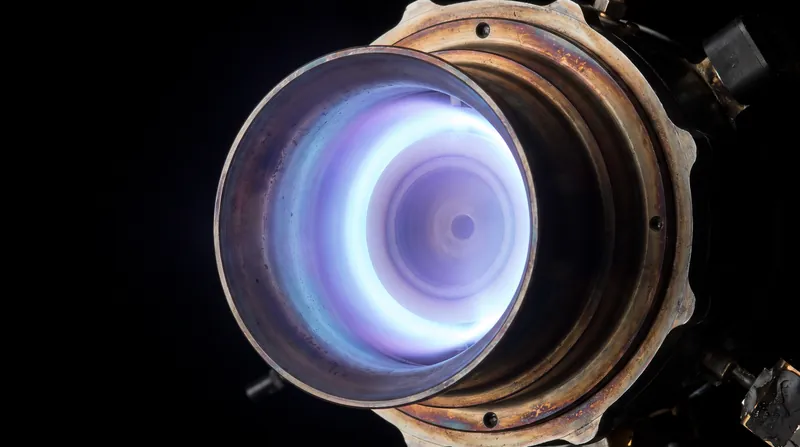 A close-up of a Hall-effect thruster operating in space, emitting a bright blue-purple plasma plume.