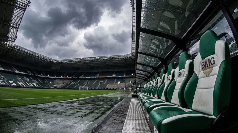 An empty managerial dugout at Borussia-Park stadium with green and white seats under overcast skies.