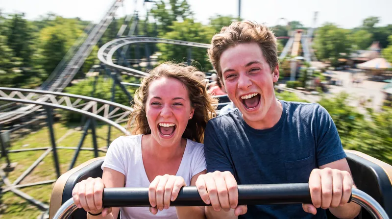 A young couple laughing and screaming while riding a fast roller coaster on a sunny day.