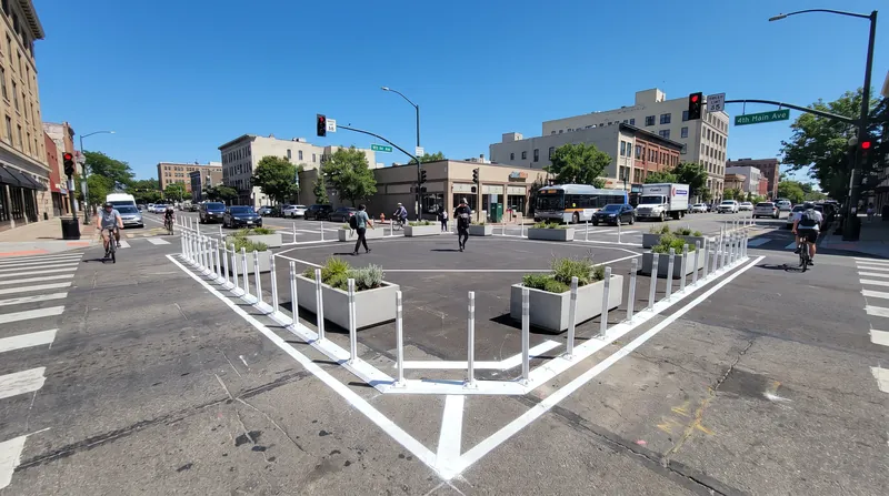 A city intersection featuring white traffic paint, flexible delineator posts, and preformed concrete planters for pedestrian safety.