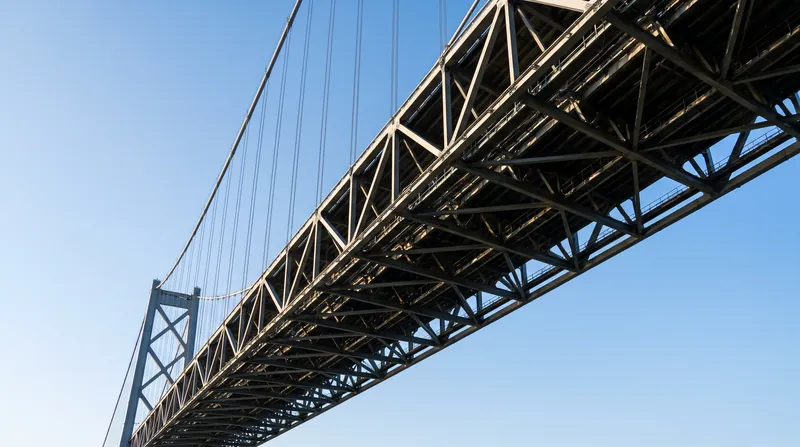 A low-angle view of a modern suspension bridge showing the complex steel lattice of its open-truss deck.