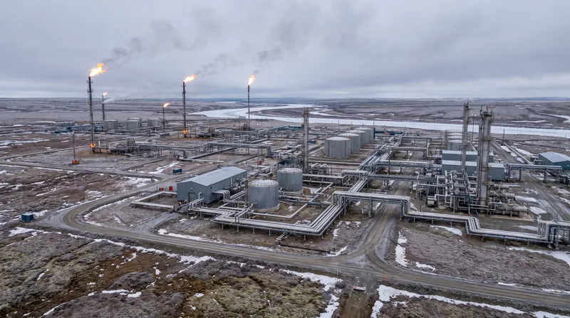 Aerial view of a large industrial gas processing facility with flare stacks and storage tanks in a tundra landscape.