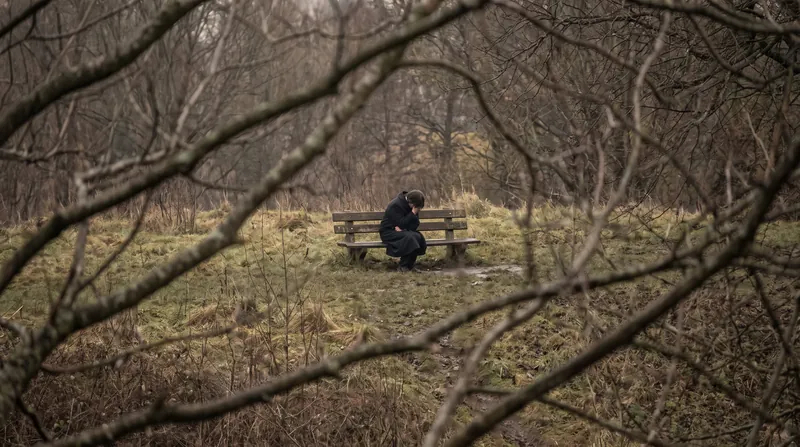 A person sitting alone on a park bench viewed through tree branches, illustrating psychological distance.