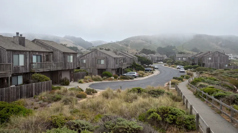 A coastal California residential neighborhood with wood-sided townhomes surrounded by coastal grasses and rolling hills.
