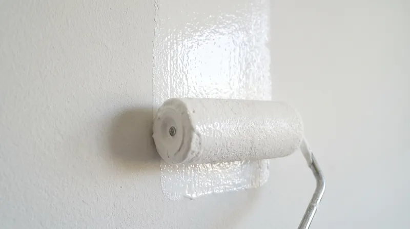 A close-up of a paint roller applying white paint to a textured interior wall.