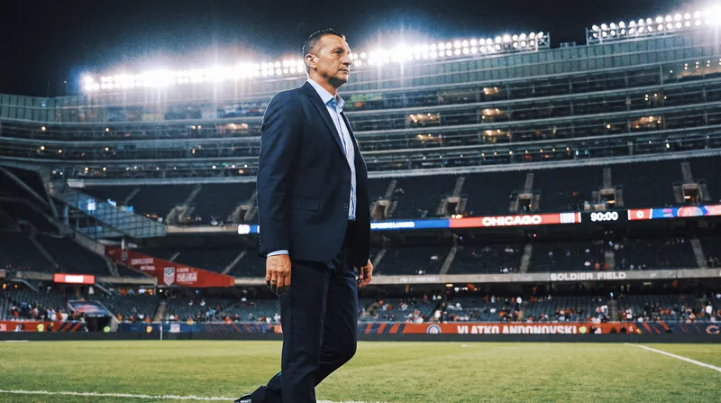 Vlatko Andonovski standing on a soccer pitch in a professional suit under bright stadium lights.