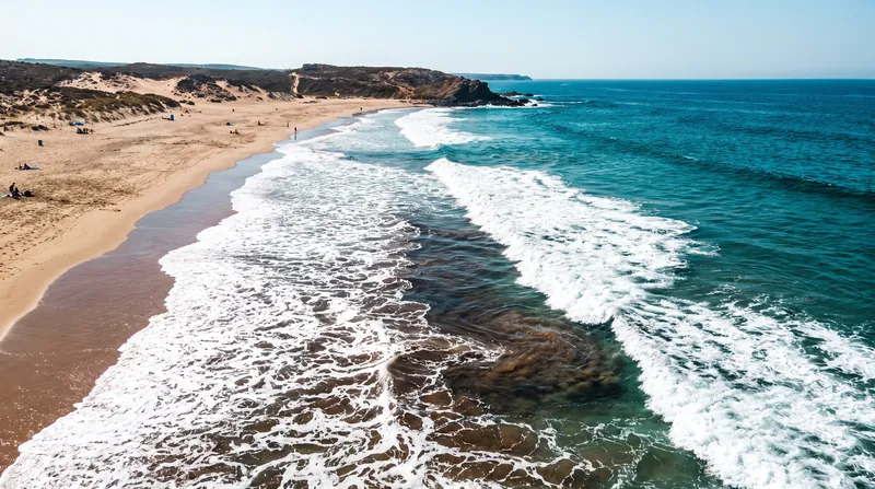 Aerial view of a beach showing a dark, narrow channel of water flowing offshore through breaking waves.