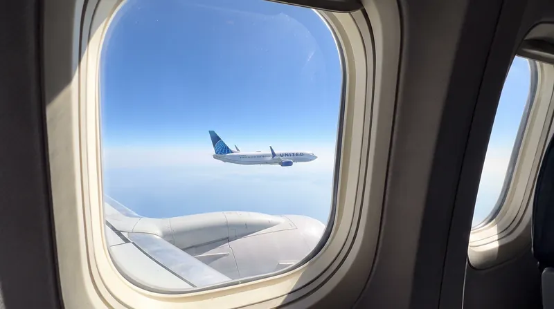 A view from an airplane window showing another commercial jet flying parallel in the distance against a blue sky.