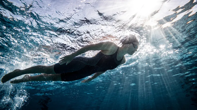 An elite swimmer glides underwater in a perfectly streamlined position, avoiding the turbulent surface waves above.