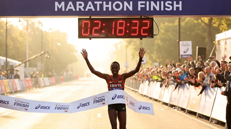 Female Kenyan runner crosses a marathon finish line with arms raised in celebration.