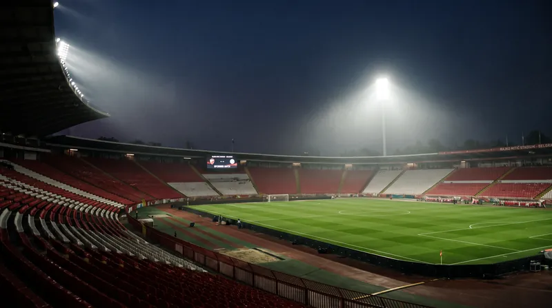 The empty green pitch and red and white seating of the Rajko Mitić Stadium in Belgrade under floodlights.