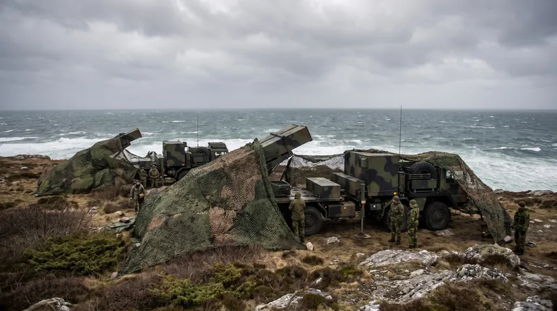 A camouflaged coastal defense cruise missile battery deployed on a rugged shoreline under an overcast sky.