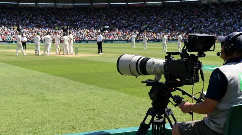 A professional television broadcast camera focused on a green cricket pitch inside a large stadium.