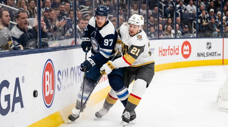 Two NHL hockey players in contrasting jerseys battling aggressively for the puck along the boards.