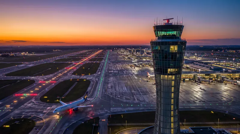 A tall air traffic control tower stands illuminated against a dusk sky overlooking a busy airport tarmac.