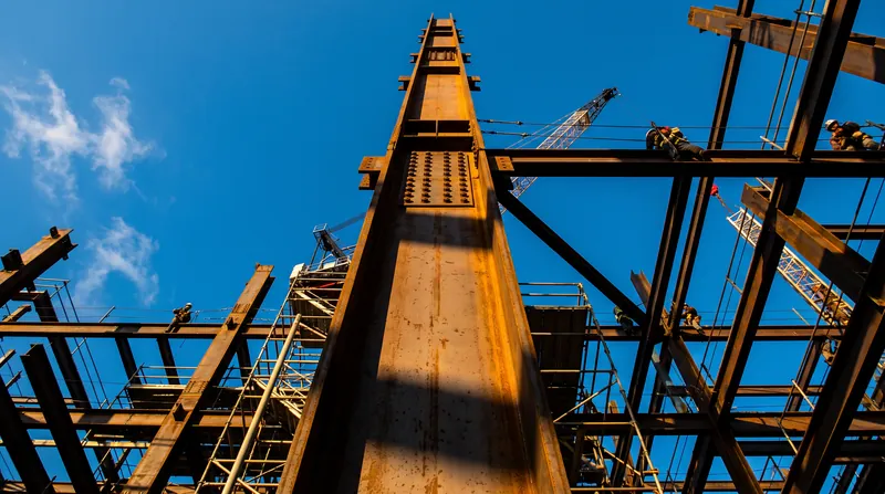 A massive steel I-beam column stands vertically under construction in a modern skyscraper against a blue sky.