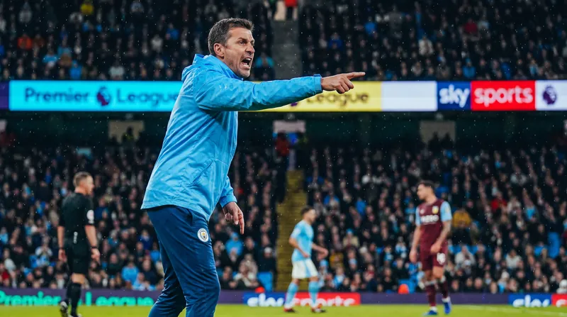 A football manager in a tracksuit stands on the touchline pointing instructions during a match.
