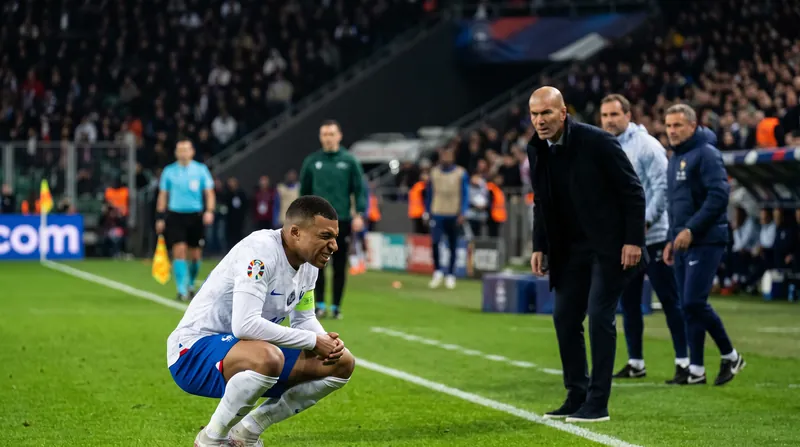 Kylian Mbappé of France holds his left knee in pain on the pitch as Zinedine Zidane watches.