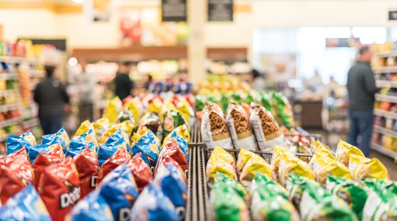 A brightly lit grocery store aisle lined with colorful bags of potato chips and savory snacks.