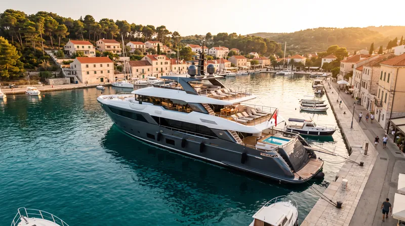 A large, multi-deck luxury superyacht moored in a sunny Mediterranean marina with clear blue water.