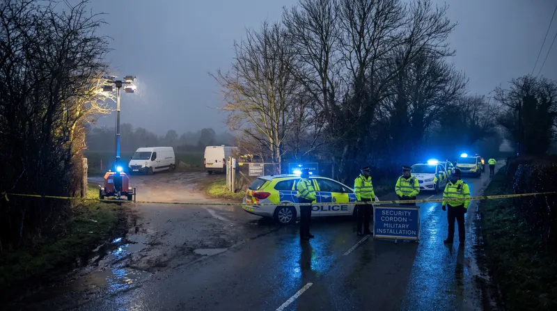A British police security cordon with flashing blue lights on a dark rural road, with white vans parked in the distance.