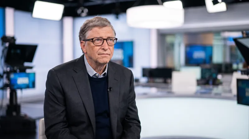 Bill Gates sitting in a television studio during an interview, wearing a dark suit.