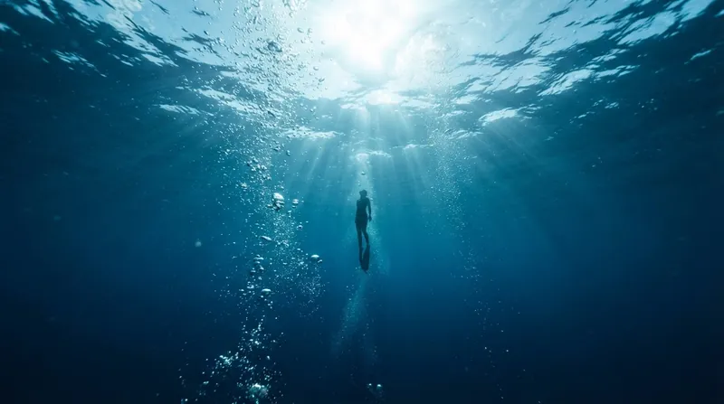 A free diver ascending toward the surface in clear blue ocean water with sun rays.