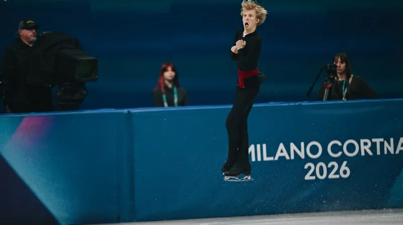 Male figure skater Daniel Grassl performing a dynamic jump mid-air during a competition with bright arena lighting.